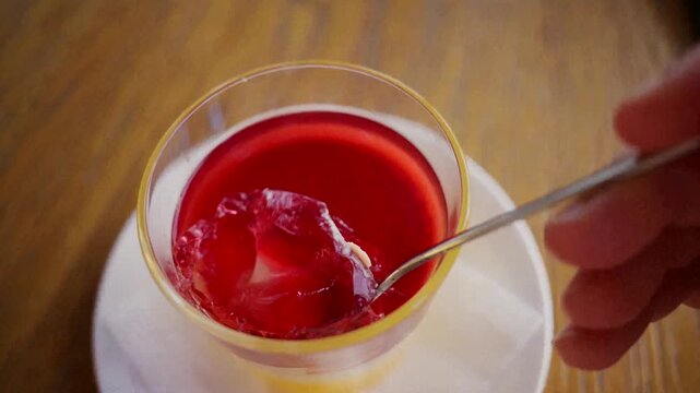 Person scooping delicious vibrant red gelatin dessert from small glass cup with metal spoon.