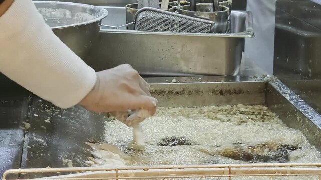 Commercial kitchen view of a chef preparing traditional tempura. Battered strips of seafood or vegetables are carefully dipped into a deep stainless steel fryer filled with hot, bubbling oil and