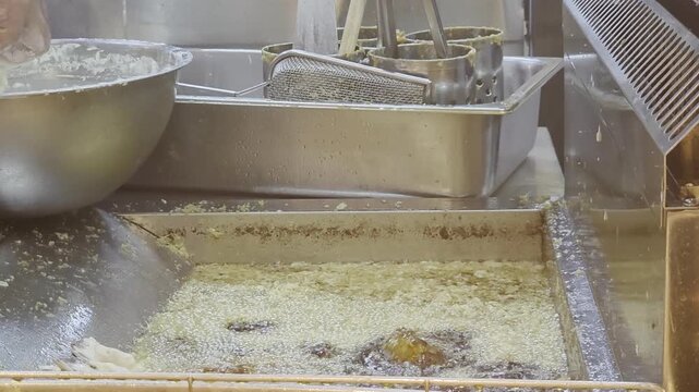 Commercial kitchen view of a chef preparing traditional tempura. Battered strips of seafood or vegetables are carefully dipped into a deep stainless steel fryer filled with hot, bubbling oil and