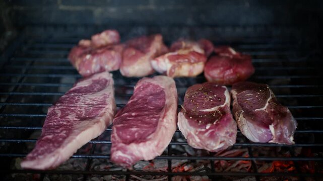 Raw sirloin steaks and marinated meat cooking on outdoor charcoal barbecue grill.