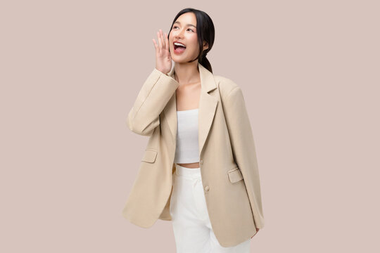 Excited young Asian businesswoman shouting or whispering secret with hand near mouth, studio portrait on beige background.