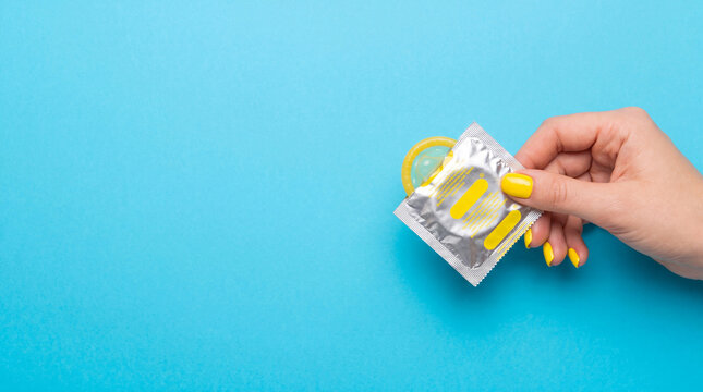 Close-up of a hand with yellow manicure holding a foil-wrapped condom against a bright blue background. Safe sex and contraception concept.