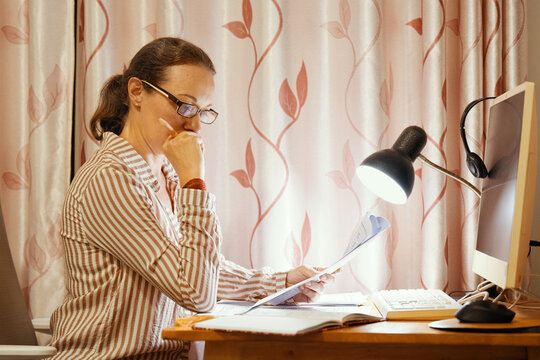 Focused middle-aged woman in glasses sitting at home desk and reading paper documents under desk lamp near computer monitor in evening.