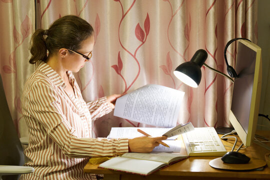 Mid-adult woman in glasses turning pages of documents at home desk with lamp and computer monitor while working on project in evening.