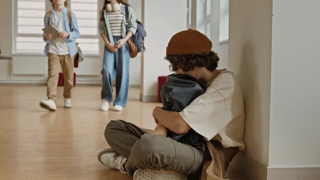 Caucasian teenager boy sitting on school hallway floor, clutching backpack while classmates walk past. Perfect for school bullying awareness, teenage mental health, or youth counseling campaigns