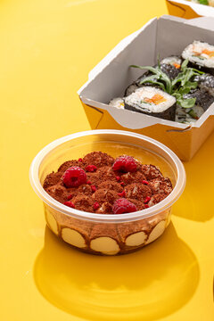 Healthy food delivery meal set with creamy tiramisu dessert cocoa powder raspberries and sushi rolls on yellow background