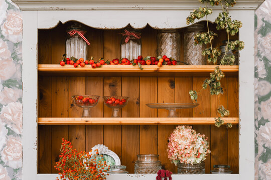A beautiful vintage wooden cabinet is decorated with glass jars, red berries, and dry autumn hydrangea flowers. Cozy rustic home interior design and seasonal fall lifestyle.