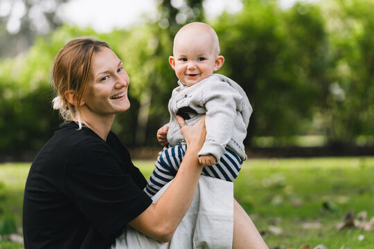 A young happy mother sits on the green park grass holding her lovely baby boy on a warm autumn day. Authentic family lifestyle representing tender parenting and outdoor recreation.