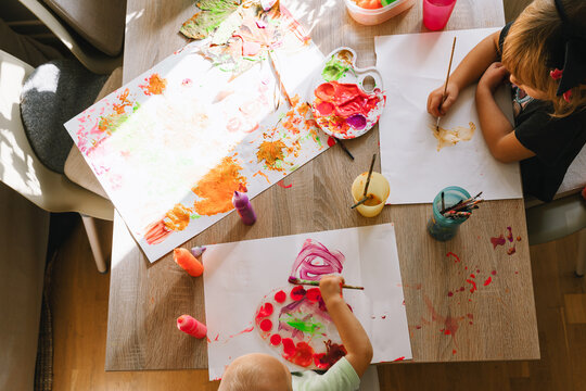 Two young children are creatively painting with bright colors on large white paper at a wooden table. Fun indoor educational activities and artistic childhood development concept.