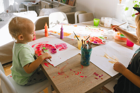 Two small preschool children sit together at a wooden table in a sunny room and paint with colorful paints. Creative early education and fun messy art activities for toddlers.
