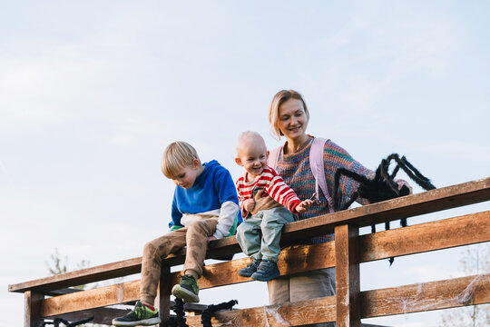 A smiling mother and two young sons sit on a rustic wooden fence playing with a large black toy spider. Joyful family Halloween preparation and fun seasonal outdoor lifestyle.