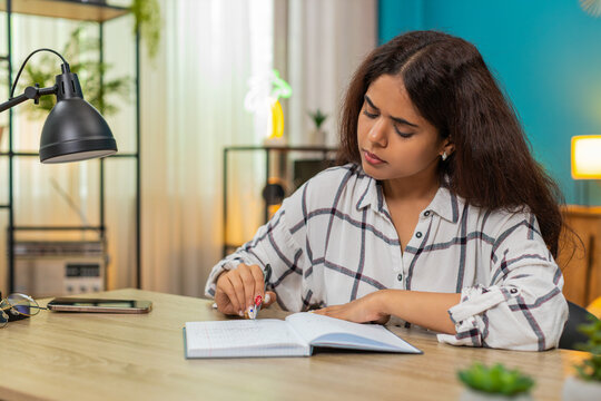 Indian woman at home writes notes in notebook then rereads lines showing careful focus and learning. Arabian girl at table studies written content reviewing details ensuring understanding and mastery.