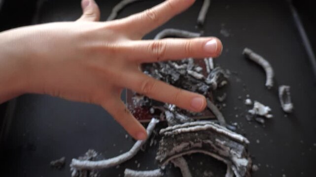 A child&rsquo;s hand stained with ash and soot after a chemical experiment. Burnt remnants of materials on a dark surface.