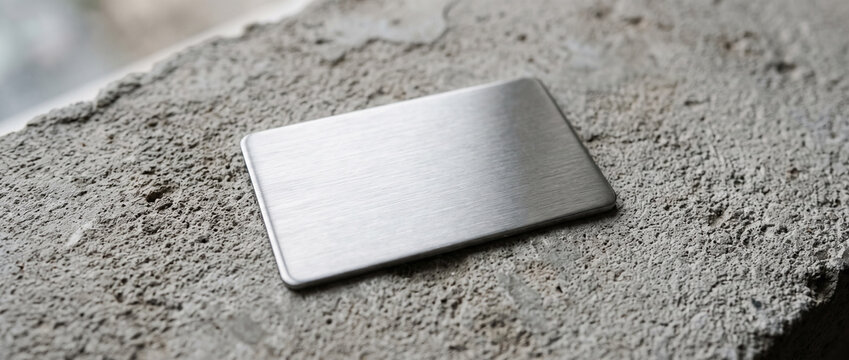 Rectangular brushed silver metal tag on rough concrete putty surface, minimalist product mockup. Close-up.