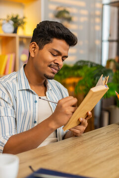 Indian man on home table reads book makes notes and pauses to think during focused study indoors at home. Arabian guy on home table reviews pages and writes ideas for learning planning and home work.