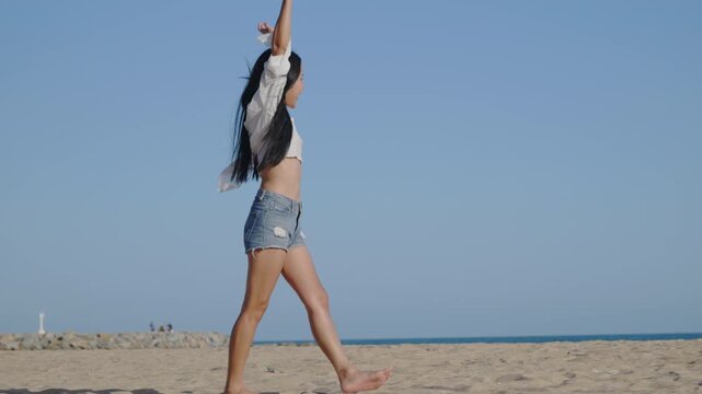 Beautiful happy lady walking on beach by ocean on tropical vacation, wearing white shirt with denim shorts and holding straw hat while smiling under clear blue sky during summer travel.
