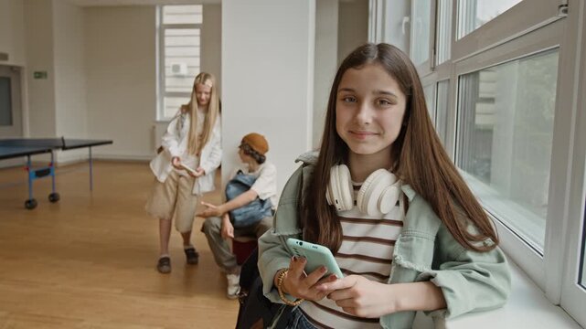 Portrait of Caucasian teenage girl holding smartphone and looking at camera in school hallway. Clip is perfect for educational apps, youth technology marketing, or social media campaigns