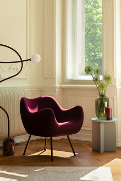 Elegant modern burgundy velvet armchair in stylish bright living room interior with sunlight, green plant in vase, and artistic floor lamp