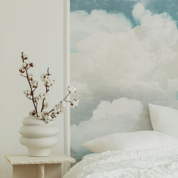 Minimalist bedroom interior design with a cloudy sky wall mural, white ceramic vase with dried cotton branch, and cozy white bed linen