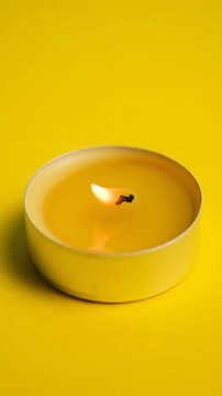 Burning tealight candle with warm flickering flame on bright yellow background, close-up minimal composition showing glowing wick, soft light, warmth, relaxation, and cozy atmosphere.