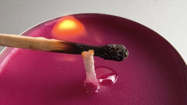 Lighting a tealight candle wick with a wooden match in extreme close-up, showing flame ignition, a burning matchstick, and melted wax during a simple candle lighting process.