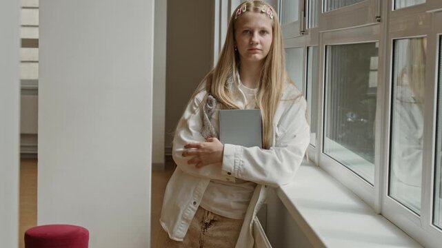 Portrait of Caucasian teenage girl standing by window in school corridor holding book and looking at camera. Footage is ideal for educational marketing and high school enrollment campaigns