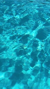 Abstract turquoise swimming pool water surface with gentle ripples, shimmering sunlight reflections, and moving wave patterns.