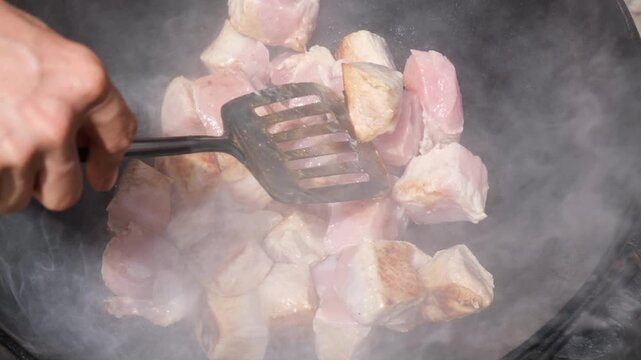 Frying large pieces of pork meat in a hot cast iron cauldron over an open fire outdoors, stirring and turning the meat with spatula.