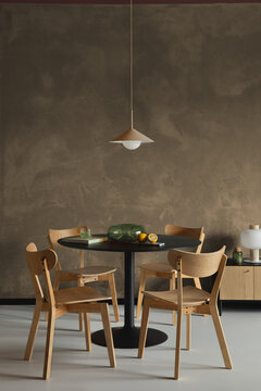 Modern dining area with wooden chairs, round black table, pendant lamp and minimalist decor against textured concrete wall.