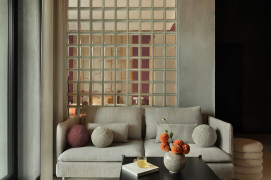 Modern minimalist living room interior with comfortable grey sofa, decorative glass block wall, and warm earthy tones creating cozy ambiance