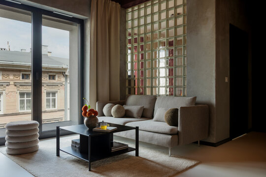 Modern minimalist living room interior with grey sofa, coffee table, glass block wall partition, and large window overlooking city architecture