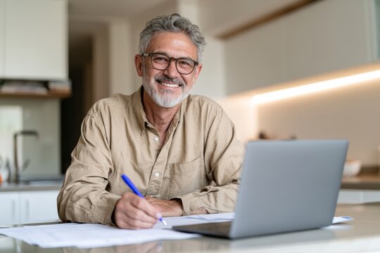 Older Man Smiling While Writing and Working on Laptop at Home