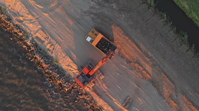 Excavator loading dump truck during dike reinforcement at sunset