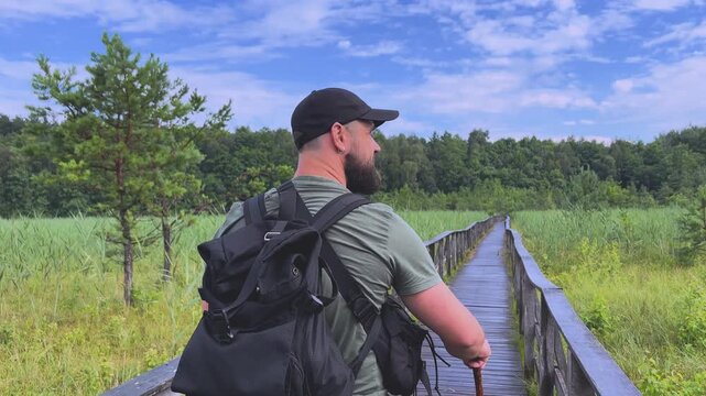 Bearded male hiker walking on a wooden boardwalk through a swamp and turning back to smile at the camera