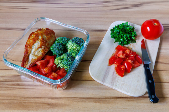 Glass container with roasted chicken breast broccoli and chopped tomatoes near wooden cutting board, healthy meal prep and fitness food concept