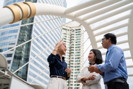 Three business professionals having discussion on outdoor pedestrian skywalk bridge with modern city skyscrapers background, corporate teamwork conversation