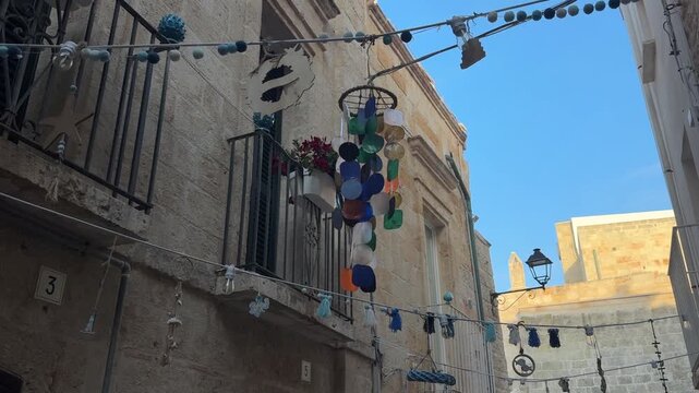 Traditional stone buildings in Monopoli old town, Italy, feature a balcony with hanging ornaments and street decorations beneath a clear blue sky. Puglia travel scene in a historic European town area.
