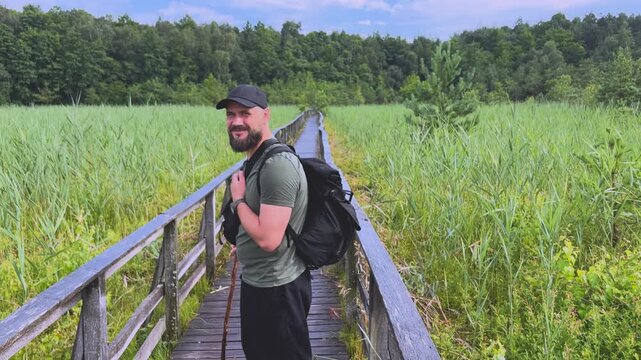 Bearded male hiker walking on a wooden boardwalk through a swamp and turning back to smile at the camera