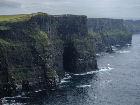 Rugged cliffs rise steeply from dark ocean waves. Green moss carpets ancient rock faces. Stormy skies loom overhead with heavy clouds. Water crashes against cliff edges, creating white foam