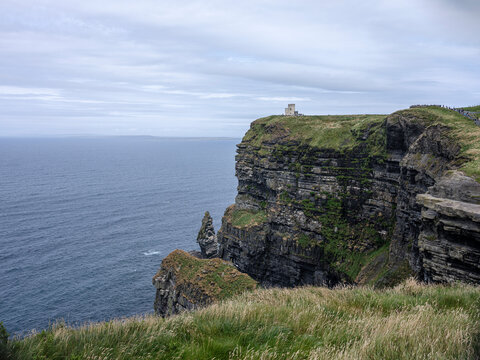 Obrien cliff towers over deep blue sea. Green moss clings to stone edges. Grass sways gently in coastal breeze. Overcast sky frames dramatic landscape. Solitary tower stands on rugged peak