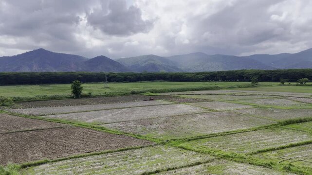 Aerial drone view of Tractor plowing a rice field with mountains beneath a cloudy sky in Southern Palawan