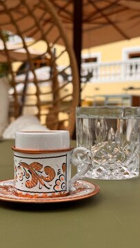 Decorative ceramic espresso cup and saucer beside a crystal water glass on a relaxed outdoor cafe table in warm light.