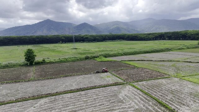 Aerial drone view of Tractor working a rice field with mountains beneath cloudy skies in Southern Palawan