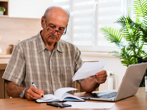 Senior man writing notes while reviewing financial documents, calculating expenses and checking his bank account on laptop at home.