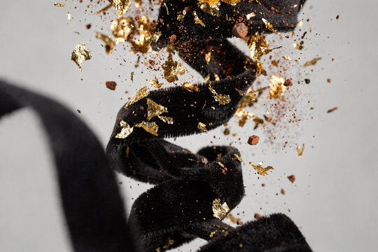 Abstract composition of twisted black velvet ribbon with flying gold leaf flakes and particles on gray background