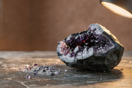 Raw purple amethyst crystal geode illuminated by a warm lamp on a dark textured surface with scattered crystal dust