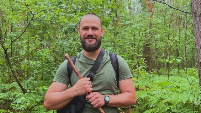 Portrait of a bearded male hiker with backpack and walking stick standing in a green forest looking at camera