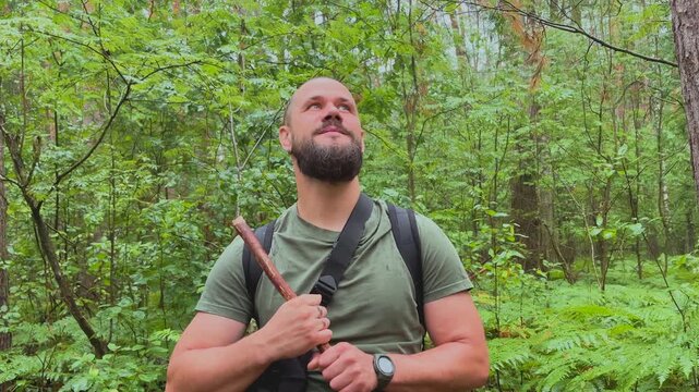 Portrait of a bearded male hiker with backpack and walking stick standing in a green forest looking at camera