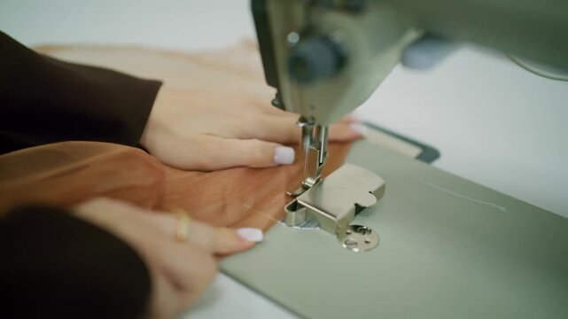 Close up of woman hands sewing brown fabric with industrial sewing machine in professional atelier.