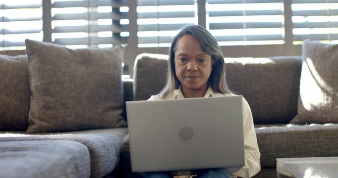 Mature African American woman positioning laptop and typing while working on sectional sofa at home